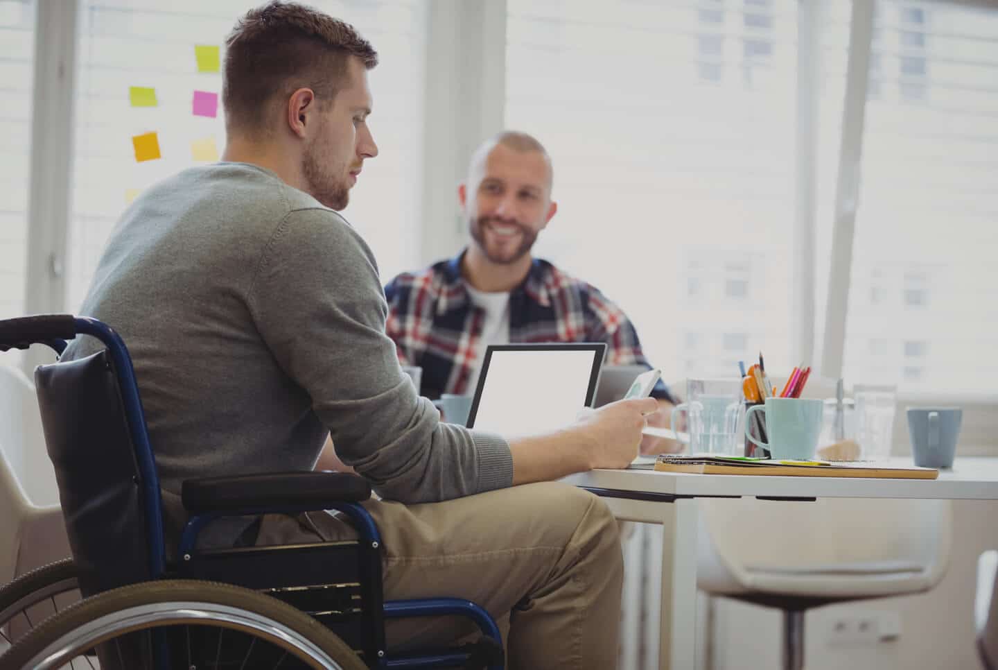 Man in wheelchair working on laptop in office with a coworker smiling